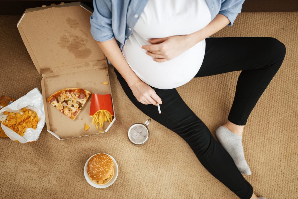Pregnant woman with belly smoking and eats unhealthy food at home. Pregnancy,